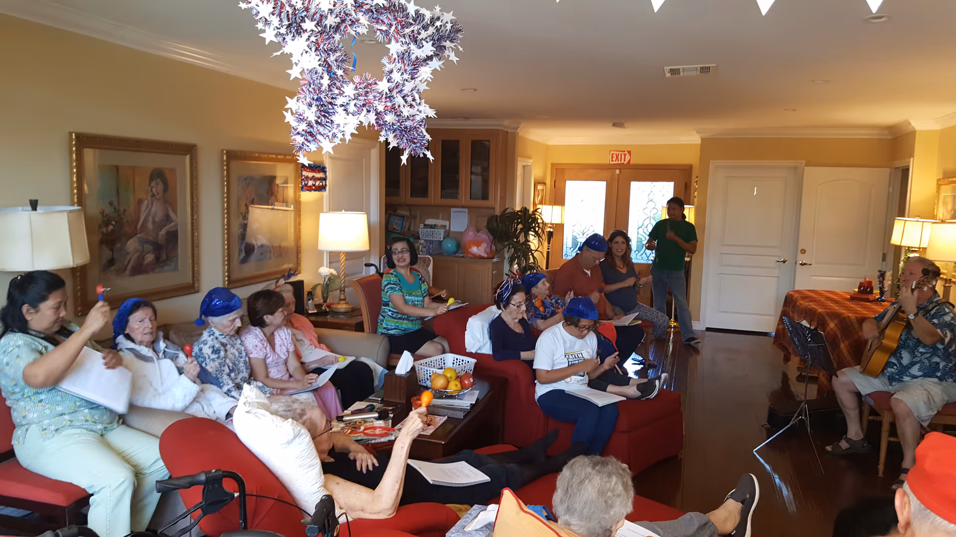 A group of residents and staff gathered in a bright living room for a musical/activity session, with one person playing guitar.