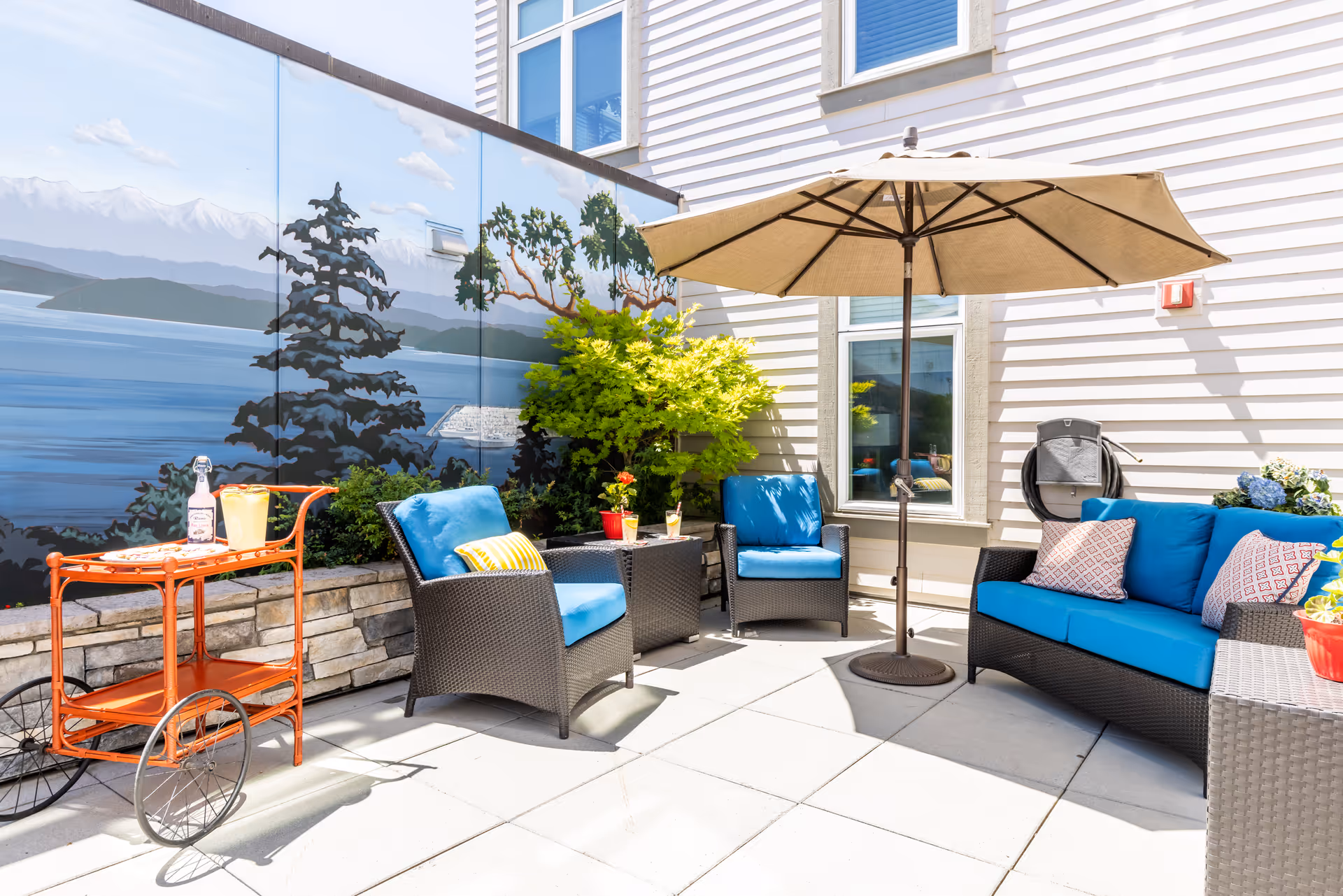 Outdoor patio area with wicker furniture including a sofa and two chairs with blue cushions, a large beige umbrella, a small table with drinks, and an orange serving cart. A mural depicting a lake and trees is painted on the wall behind the seating area.