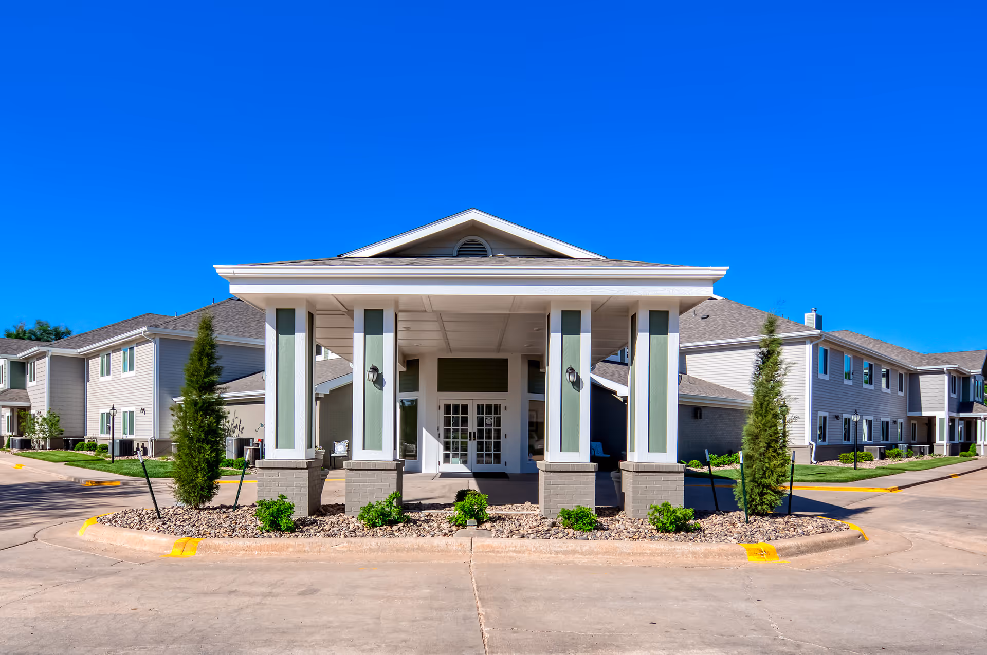 Front exterior view of a senior living facility named Parkwood Village, featuring a covered entrance with four columns, landscaped greenery, and clear blue sky.