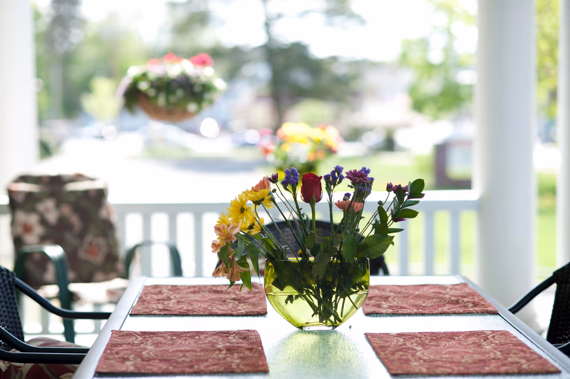 A glass table with four red patterned placemats and a green glass vase holding a colorful bouquet of flowers, situated on a porch with white railing and outdoor chairs with floral cushions. The background shows a blurred view of greenery and hanging flower baskets.