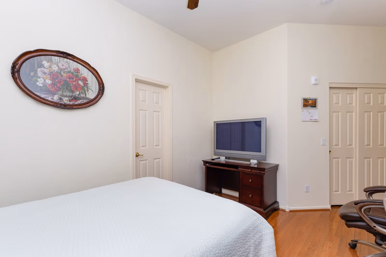 A bedroom with a white bedspread, a wooden TV stand with a flat-screen TV, a floral painting in an oval wooden frame on the wall, a closed door, a calendar on the wall, and a brown office chair on hardwood flooring.