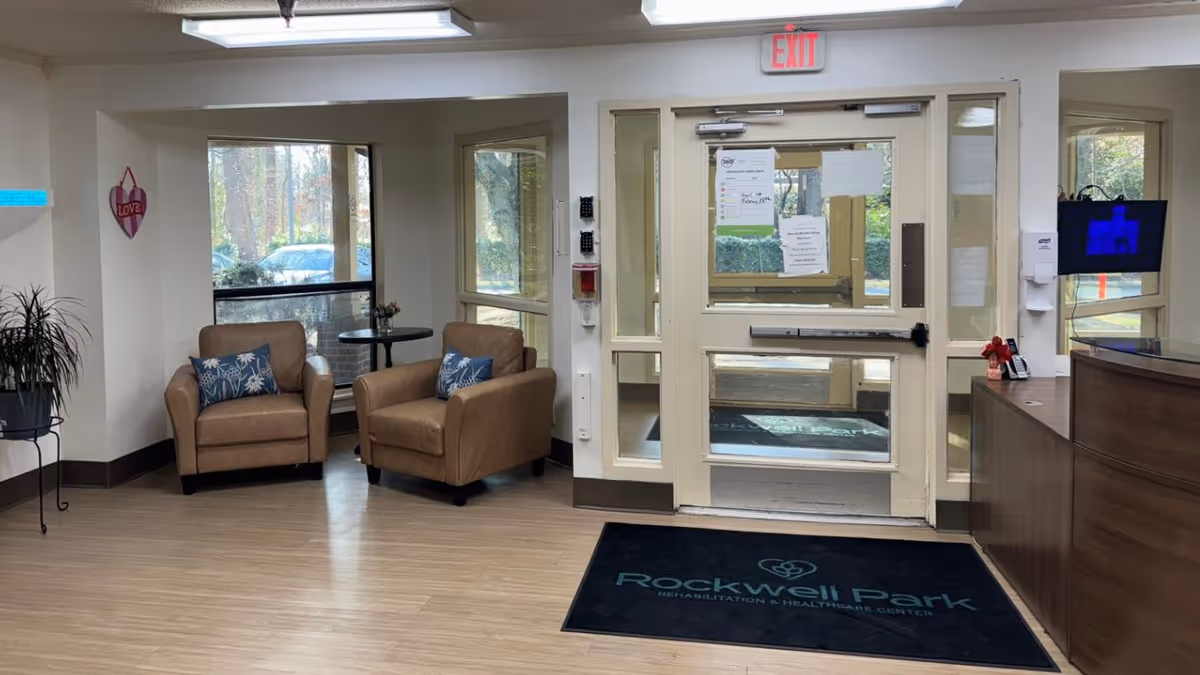 Entrance area of Rockwell Park Rehabilitation and Healthcare Center featuring a glass door with exit sign above it, two brown armchairs with blue patterned pillows near a window, a small round table with a flower vase, a plant on a stand, and a reception desk with a computer monitor on the right side. A black floor mat with the facility's name is placed in front of the door.