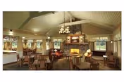 A spacious dining room with multiple wooden tables and chairs arranged neatly. The room features a high vaulted ceiling with exposed beams, large windows allowing natural light, and a stone fireplace at the center with a mounted TV above it. Warm lighting fixtures hang from the ceiling, creating a cozy atmosphere.