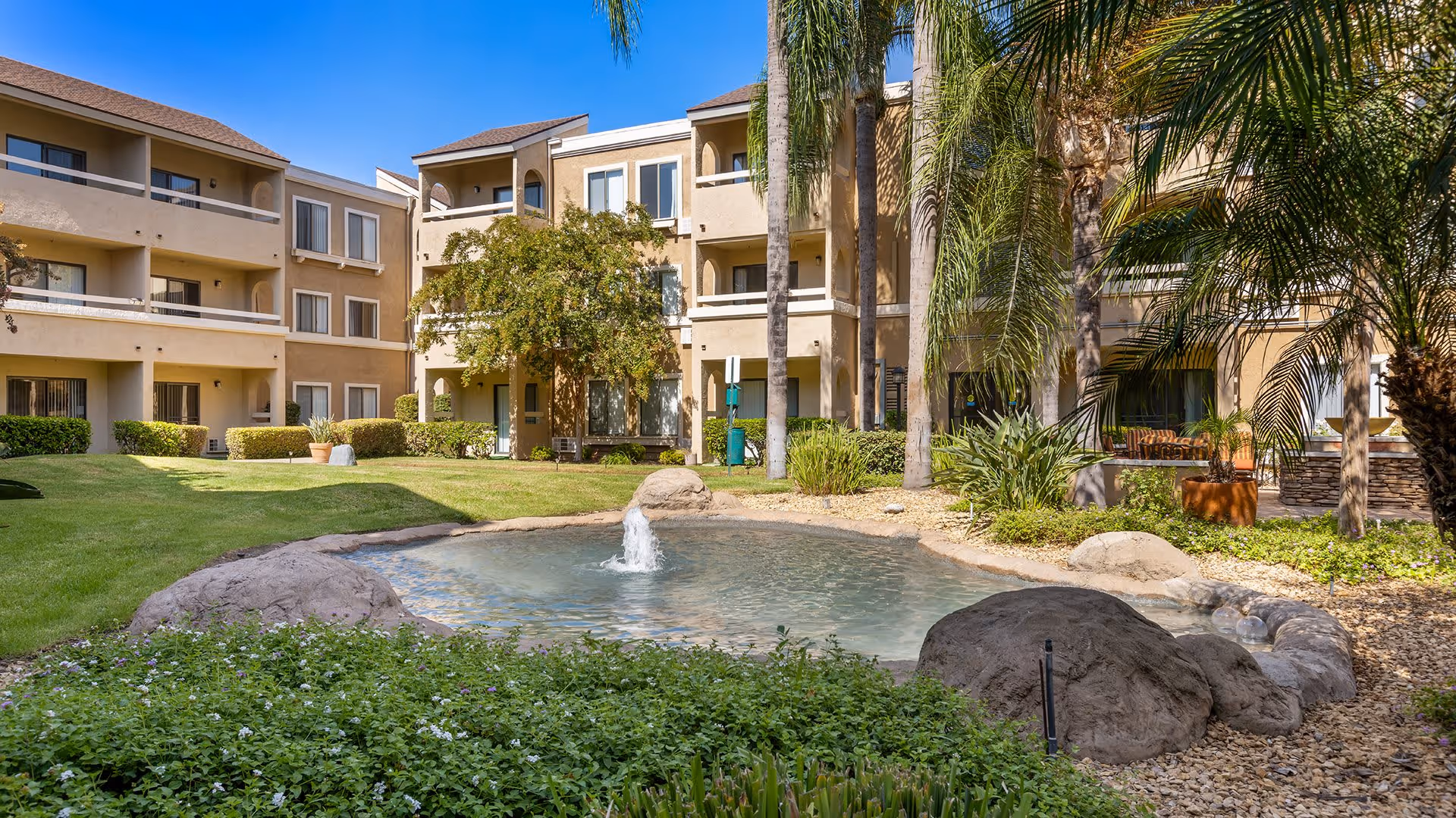 Outdoor courtyard area of a senior living facility with a small pond featuring a water fountain, surrounded by rocks, green grass, palm trees, and shrubs. Beige multi-story buildings with balconies and windows are visible in the background under a clear blue sky.