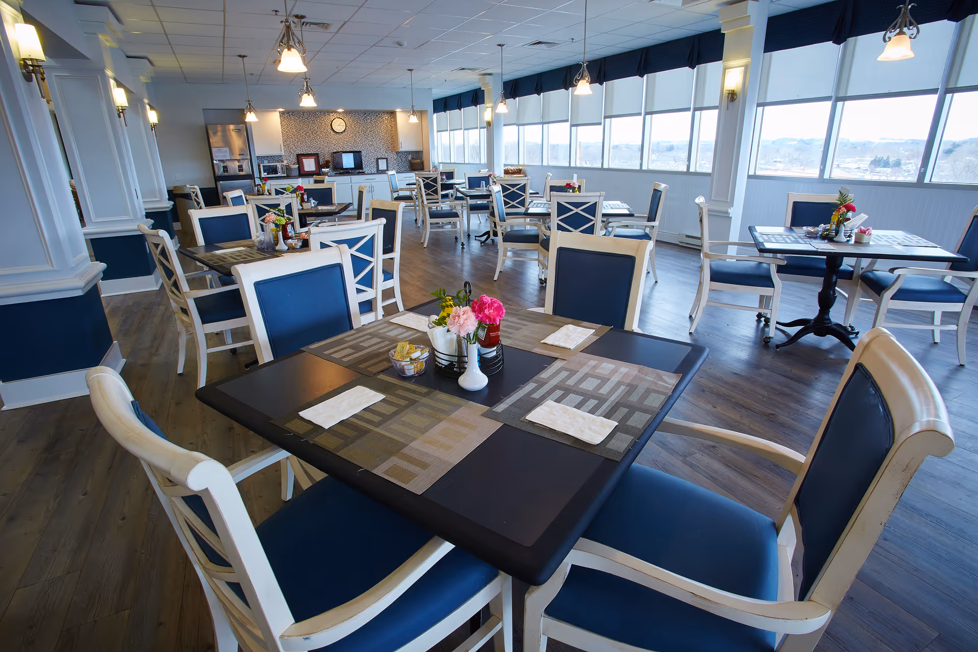 A bright dining room with multiple tables and chairs arranged neatly. Each table has placemats, napkins, and small flower arrangements. Large windows line one side of the room, allowing natural light to fill the space. The room features wood flooring and a clean, modern design.