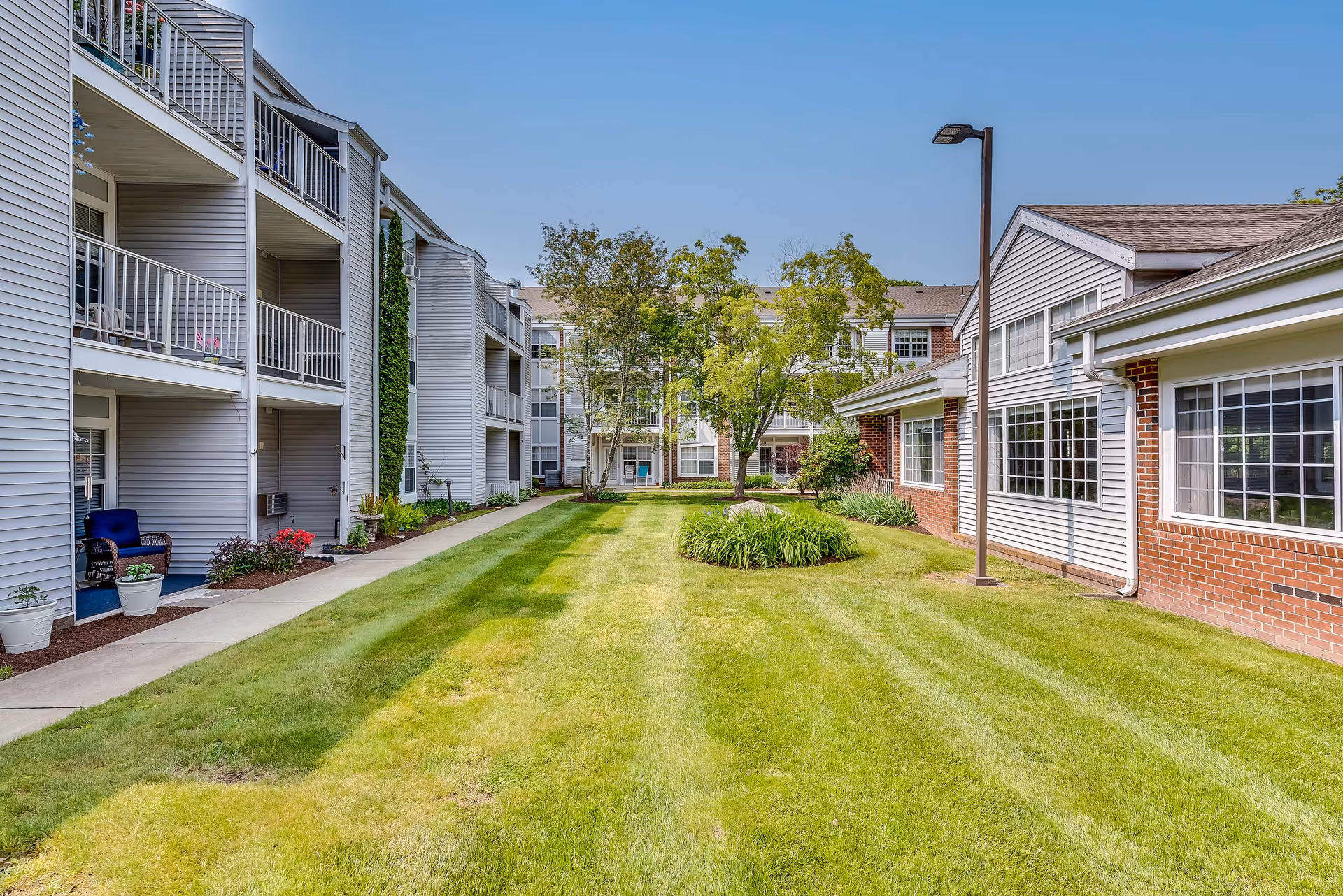 Outdoor courtyard area of Solstice Senior Living at Groton featuring a well-maintained lawn, a few trees, and shrubs. The courtyard is surrounded by multi-story residential buildings with balconies and large windows. A paved walkway runs along the left side of the grass, and a street lamp is visible on the right side near the building.