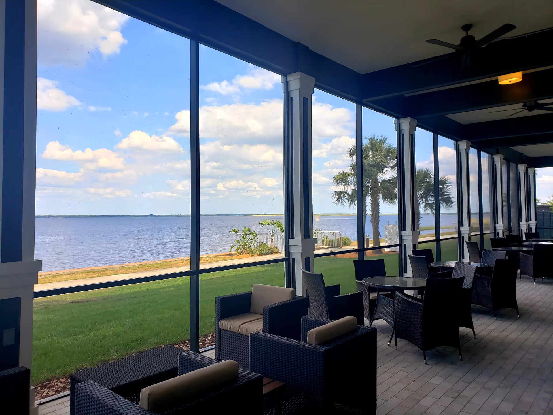 A covered outdoor patio area with wicker chairs and tables overlooking a waterfront with palm trees and a partly cloudy sky.