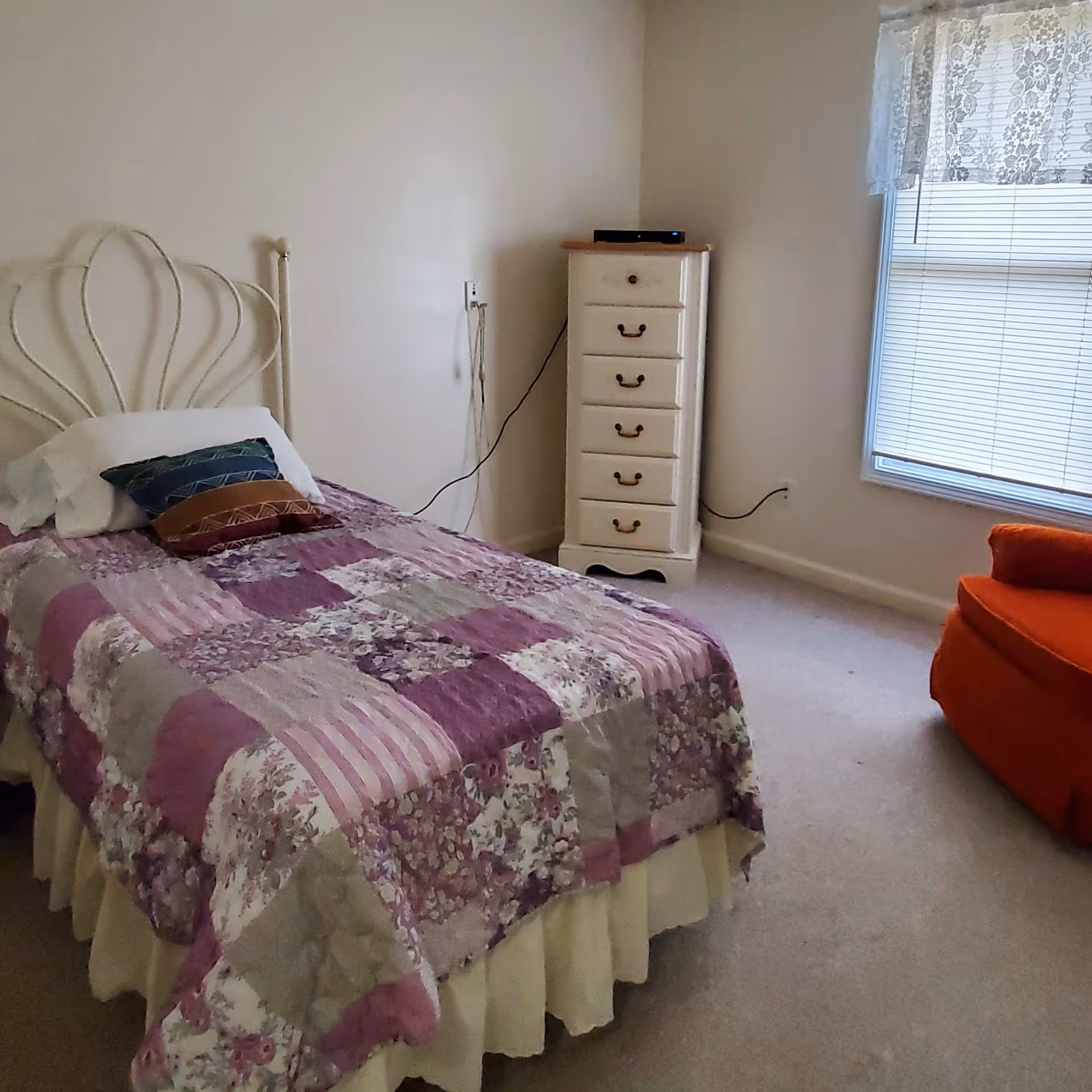 A small bedroom with a single bed covered in a purple and white patchwork quilt, a white pillow with two decorative pillows, a white chest of drawers in the corner, a window with blinds and lace curtains, and an orange armchair.