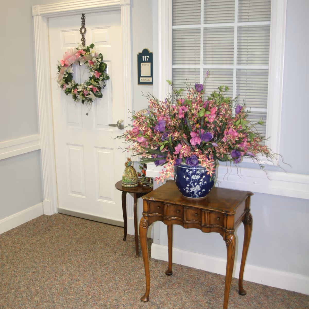 Interior hallway with a white door marked 117 decorated with a floral wreath and a wooden table holding a large blue vase of purple and pink flowers.