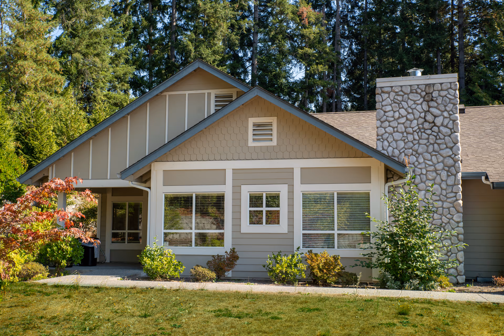 Exterior view of a single-story building with beige siding, white trim, and a stone chimney. The building is surrounded by green shrubs and trees, with a grassy lawn in the foreground under a clear sky.