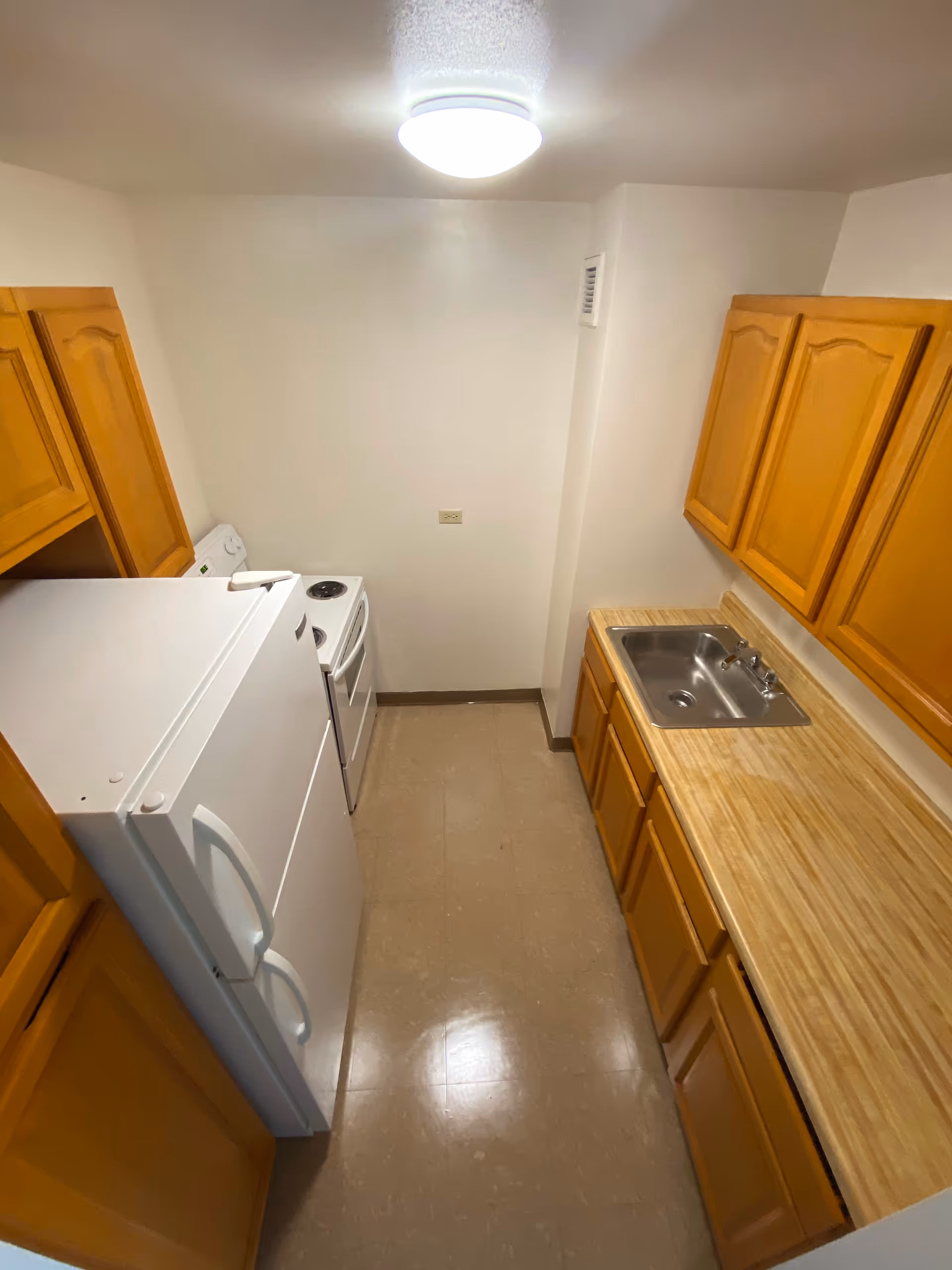 Small kitchen with wooden cabinets, a white refrigerator, a white electric stove, a stainless steel sink, and beige countertops and floor tiles under a ceiling light.