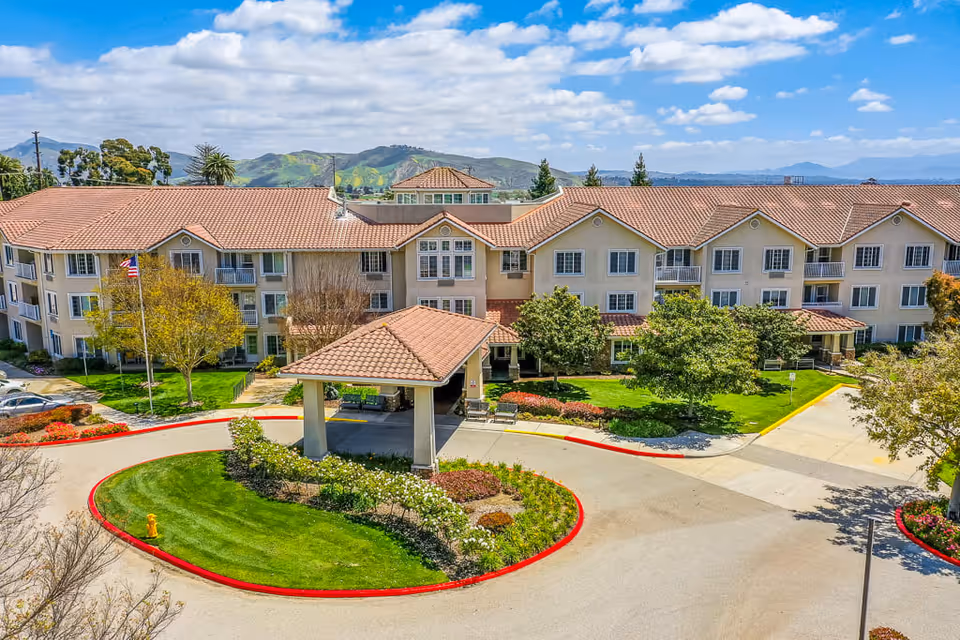 Exterior view of The Orchard Senior Living facility showing a large three-story building with beige walls and red-tiled roofs. The building is surrounded by well-maintained landscaping including green lawns, trees, and flower beds. A covered entrance with a red-tiled roof is visible in the center, with a circular driveway and a fire hydrant on a grassy island. Hills and a partly cloudy sky are in the background.