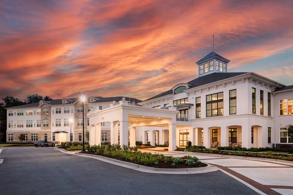 Exterior view of The Sheridan at Eastside senior living facility at sunset, featuring a large white building with multiple windows, a covered entrance, landscaped greenery, and a dramatic colorful sky with orange and pink clouds.