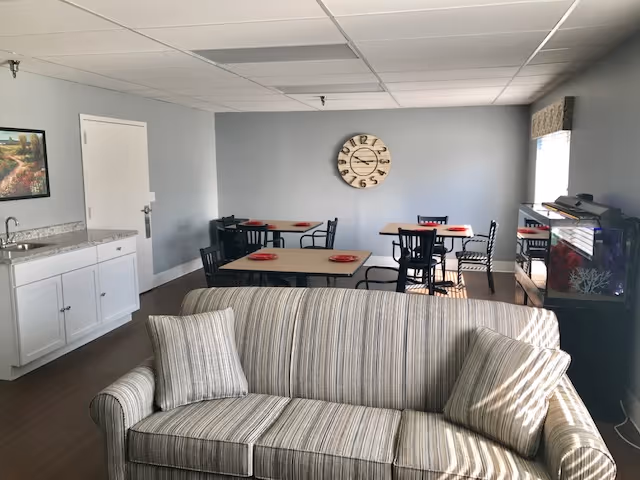 A common area in a senior living facility featuring a striped sofa with two matching pillows in the foreground. Behind the sofa are several tables with black chairs, each table set with red plates. A large clock hangs on a light gray wall, and a window with a valance allows natural light into the room. A white cabinet with a sink is on the left side, and an aquarium is visible on the right side near the window.