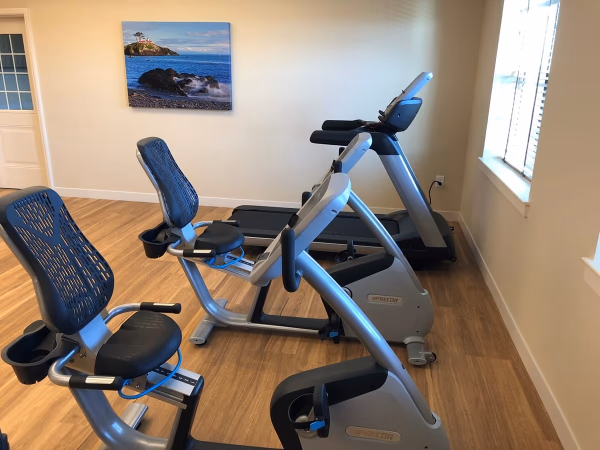 A small exercise room with two recumbent stationary bikes and one treadmill. The room has light-colored walls, wood flooring, a window with blinds, and a framed picture of a coastal scene with a lighthouse on the wall.