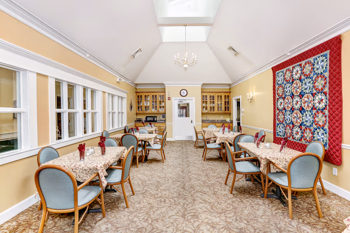 A dining room with several tables covered in patterned tablecloths, each set with folded red napkins and small candle holders. The room has beige walls, a patterned carpet, a high ceiling with a chandelier, and a large colorful quilt hanging on one wall. There are multiple windows on one side and wooden cabinets with glass doors at the far end.