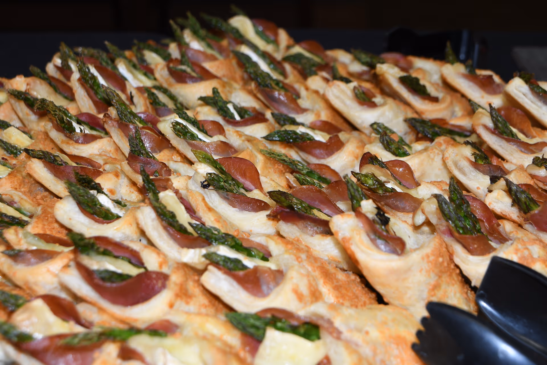 Close-up view of multiple puff pastry appetizers topped with asparagus and a slice of cured meat, arranged neatly on a tray.