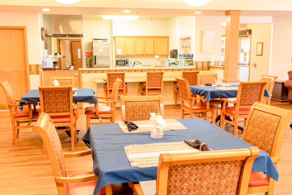 Dining area with several wooden tables covered with blue tablecloths and matching wooden chairs with patterned upholstery. The background shows a kitchen area with wooden cabinets, a refrigerator, microwave, and counter seating. The room has warm lighting and wooden flooring.