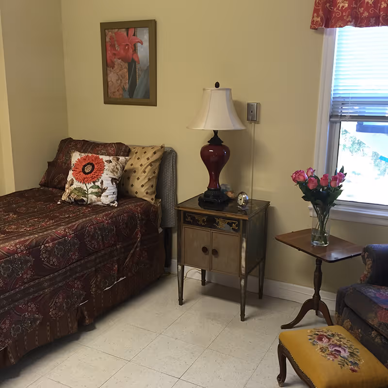 A cozy bedroom with a single bed covered in a patterned maroon bedspread and decorative pillows, a small vintage nightstand with a red lamp and a clock, a wooden side table with a vase of pink roses, a floral upholstered chair, and a window with blinds and a red valance.