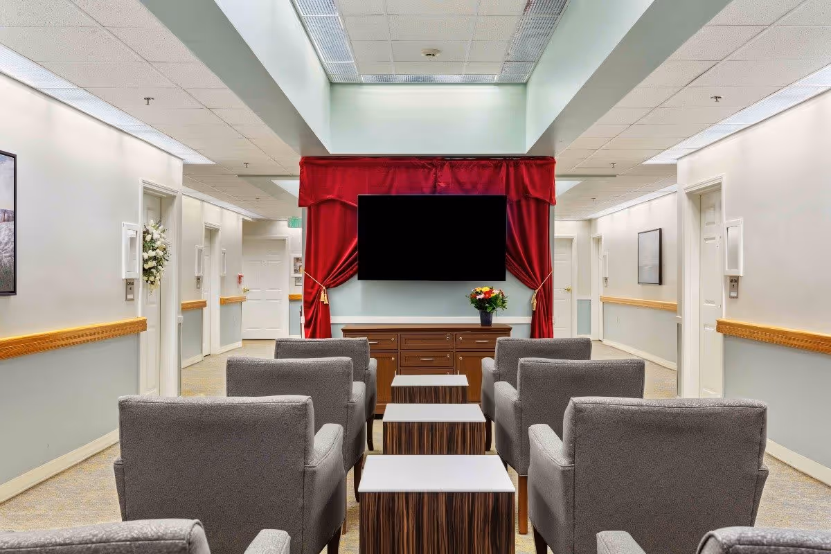 A hallway seating area in a senior living facility with six gray armchairs arranged in two rows facing a flat-screen TV mounted on a wall with red curtains. There are three small tables between the chairs, a wooden cabinet under the TV with a flower vase on top, and doors along the hallway walls.
