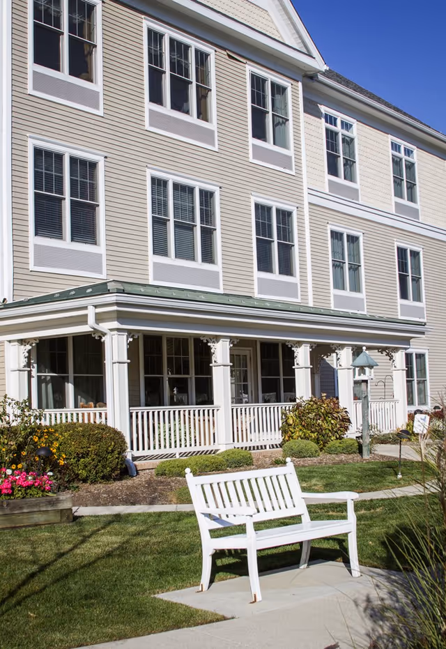 Exterior view of a multi-story senior living facility with beige siding and white trim. The building features multiple windows and a covered porch with white railings. In the foreground, there is a white wooden bench on a concrete pad surrounded by green grass, bushes, and colorful flowers under a clear blue sky.