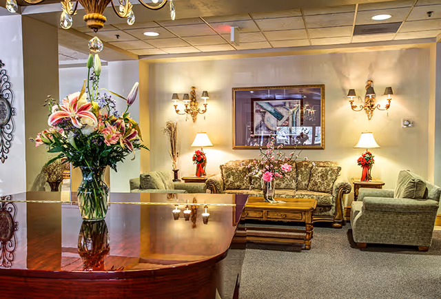 Well-lit senior living lounge with a polished wood table holding a vase of flowers and upholstered sofas, armchairs, lamps, and a coffee table in the background.