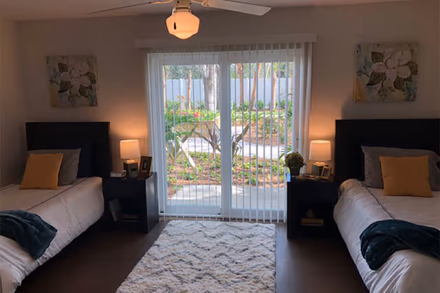 A bedroom with two single beds placed on either side of a sliding glass door with vertical blinds. Each bed has a yellow pillow and a dark green throw blanket. There are two nightstands, each with a lamp and decorative items. A ceiling fan with a light is above, and a patterned rug is on the floor between the beds. Outside the glass door, a garden with plants and a fence is visible.
