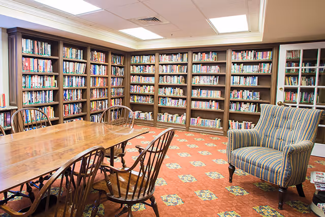 A cozy library room with wooden bookshelves filled with books lining the walls. There is a large wooden table surrounded by wooden chairs in the center of the room, and a striped upholstered armchair is positioned near the right side. The floor is covered with a patterned carpet, and the ceiling has recessed lighting.