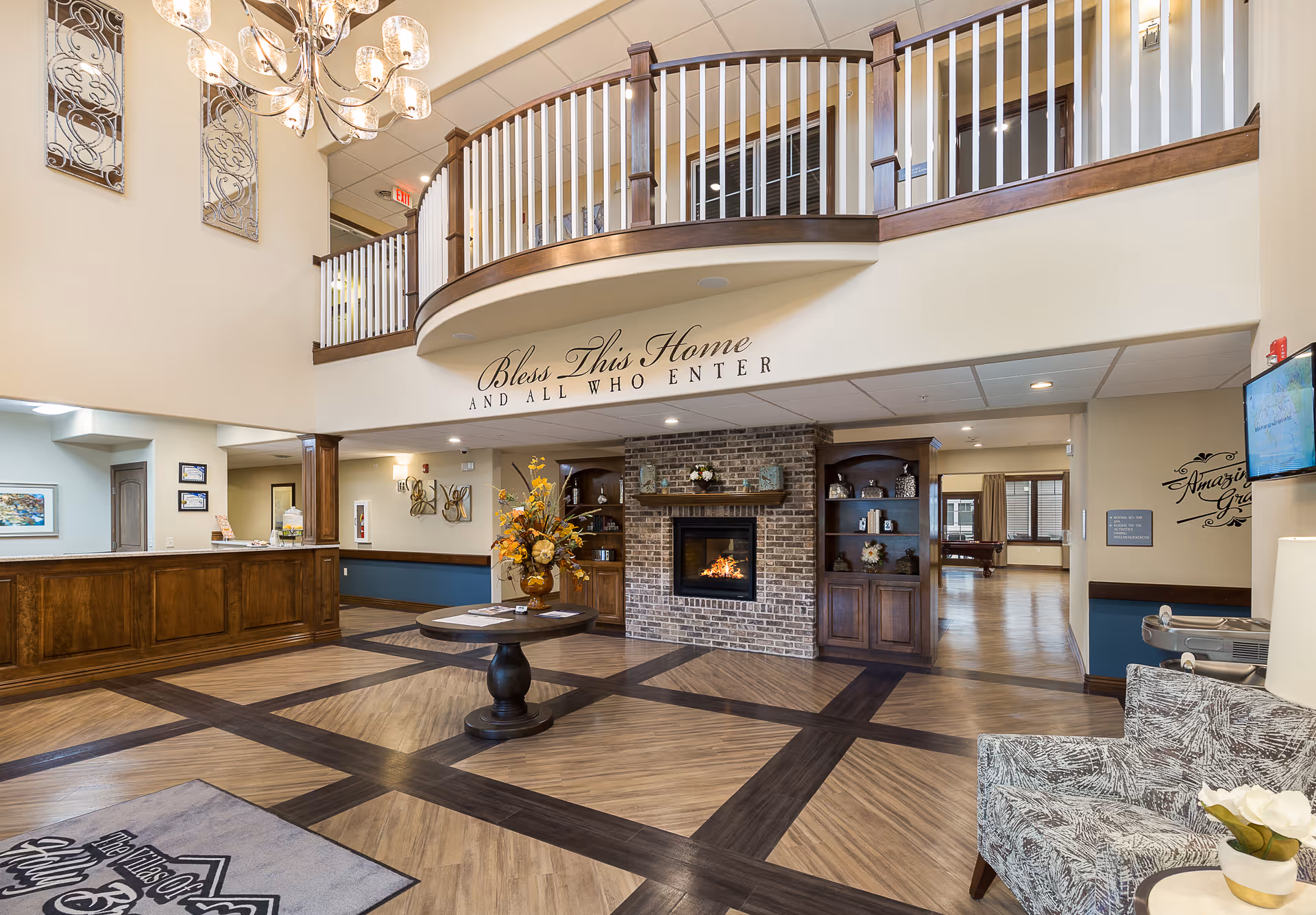 Spacious and warmly decorated assisted living facility lobby with a wooden reception desk on the left, a round table with a floral arrangement in the center, and a brick fireplace with built-in wooden shelves on either side. Above the fireplace is a sign that reads 'Bless This Home AND ALL WHO ENTER.' The area features a high ceiling with a chandelier, a balcony railing on the upper level, and comfortable seating on the right side.