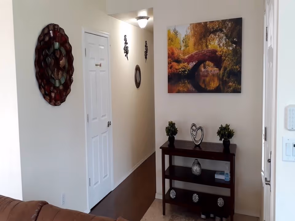 Interior view of a hallway in a senior living facility with a white door, a small dark wooden table holding decorative items including two small plants and a heart-shaped ornament, a large colorful painting of a bridge over water on the wall, and a round decorative mirror on the adjacent wall.