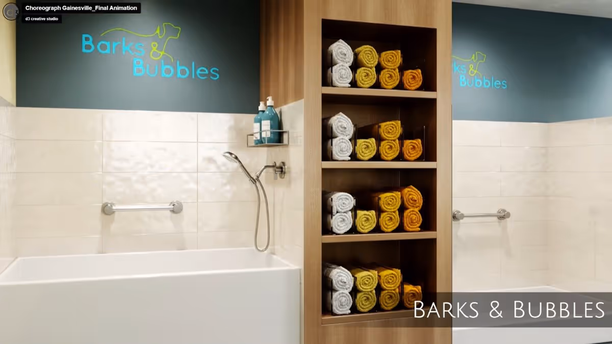 Interior view of a bathing area with two white bathtubs separated by a wooden shelving unit filled with rolled towels in white, yellow, and orange colors. The walls are tiled in white with a blue upper section featuring the text 'Barks & Bubbles' and a simple dog outline logo. Each bathtub has a handheld showerhead and a grab bar.