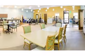 A spacious, well-lit common area in a senior living facility featuring several tables and chairs, exercise equipment, and large windows allowing natural light to fill the room. The walls are painted yellow and white, and the ceiling has modern light fixtures.