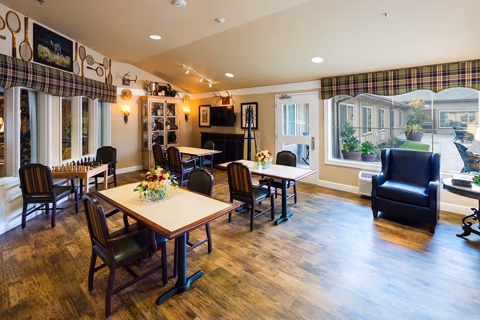 A bright and cozy common area in a memory care community featuring several tables with chairs, floral centerpieces, a chessboard on a side table, a large window overlooking an outdoor patio with plants, a comfortable armchair, and decorative wall accents including vintage tennis rackets and mounted antlers.