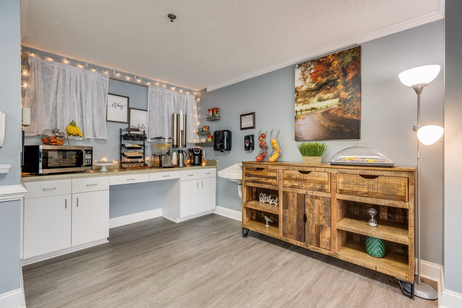 A cozy kitchenette area in an assisted living facility with white cabinets, a microwave, a coffee maker, beverage dispensers, and a wooden sideboard. The walls are painted light blue with sheer white curtains and string lights above the counter. Decorative items include a framed autumn landscape picture, fruit containers, and small decorative signs.