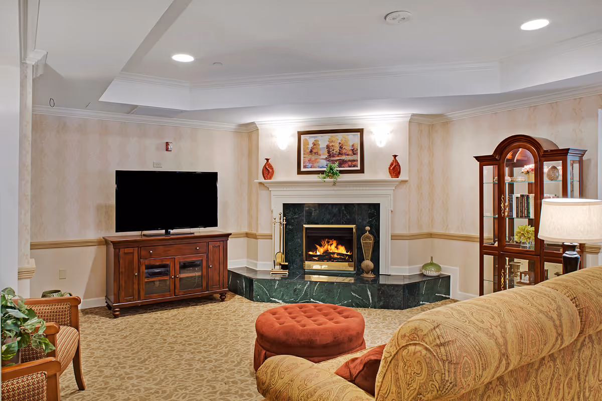A cozy living room with a patterned beige carpet, a large flat-screen TV on a wooden cabinet, a green marble fireplace with a fire burning, and a framed painting above it. There is a round red ottoman in front of a patterned sofa, a wooden display cabinet with glass doors, and a table lamp on the right side.