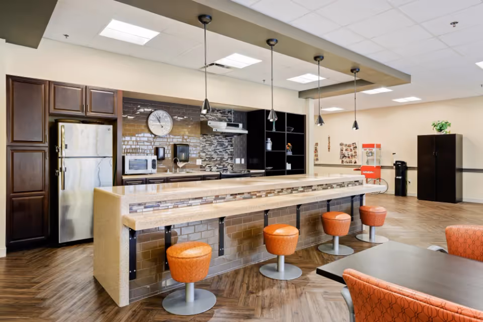A modern kitchen area in a senior living facility with a long counter and five orange swivel stools. The kitchen features dark wood cabinets, a stainless steel refrigerator, a microwave, a clock on a tiled backsplash, and three pendant lights hanging from the ceiling. In the background, there is a popcorn machine, a black cabinet, and a table with orange patterned chairs.