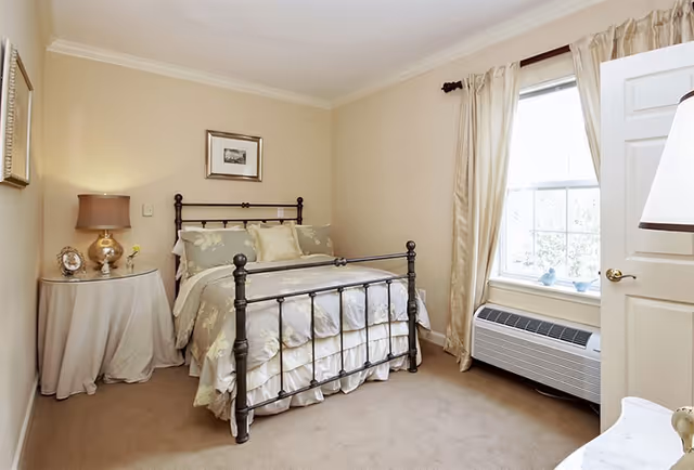 A cozy bedroom with a black metal bed frame, floral bedding, and multiple pillows. There is a round side table covered with a white cloth, holding a lamp, a framed photo, and a small flower vase. The room has beige walls, a window with cream-colored curtains, and a white door partially open. Below the window is a white air conditioning unit.