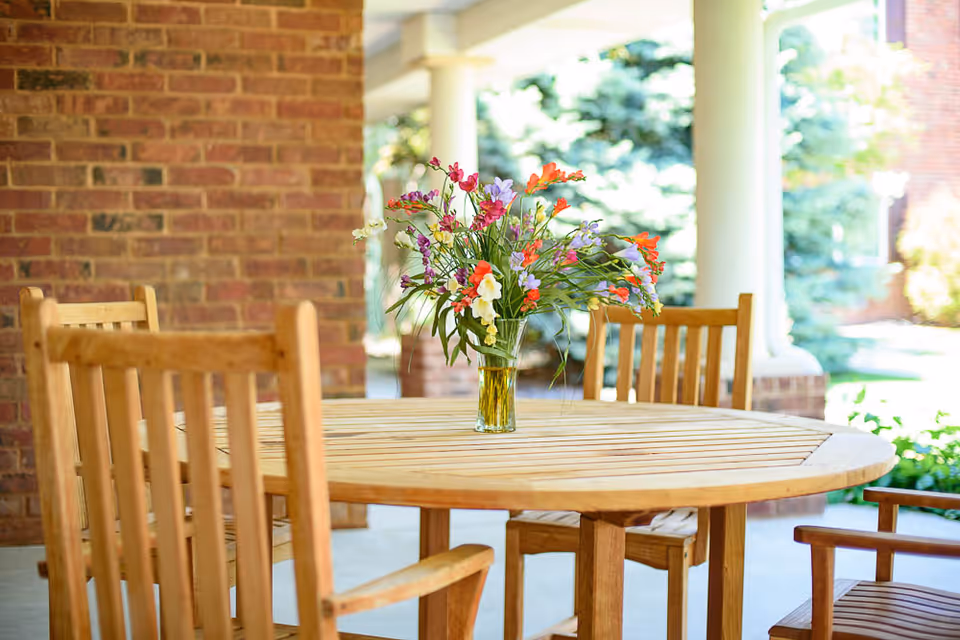 A round wooden outdoor table with a glass vase holding a colorful bouquet of flowers. The table is surrounded by wooden chairs and is set on a covered patio with brick walls and white columns. Green trees and shrubs are visible in the background.