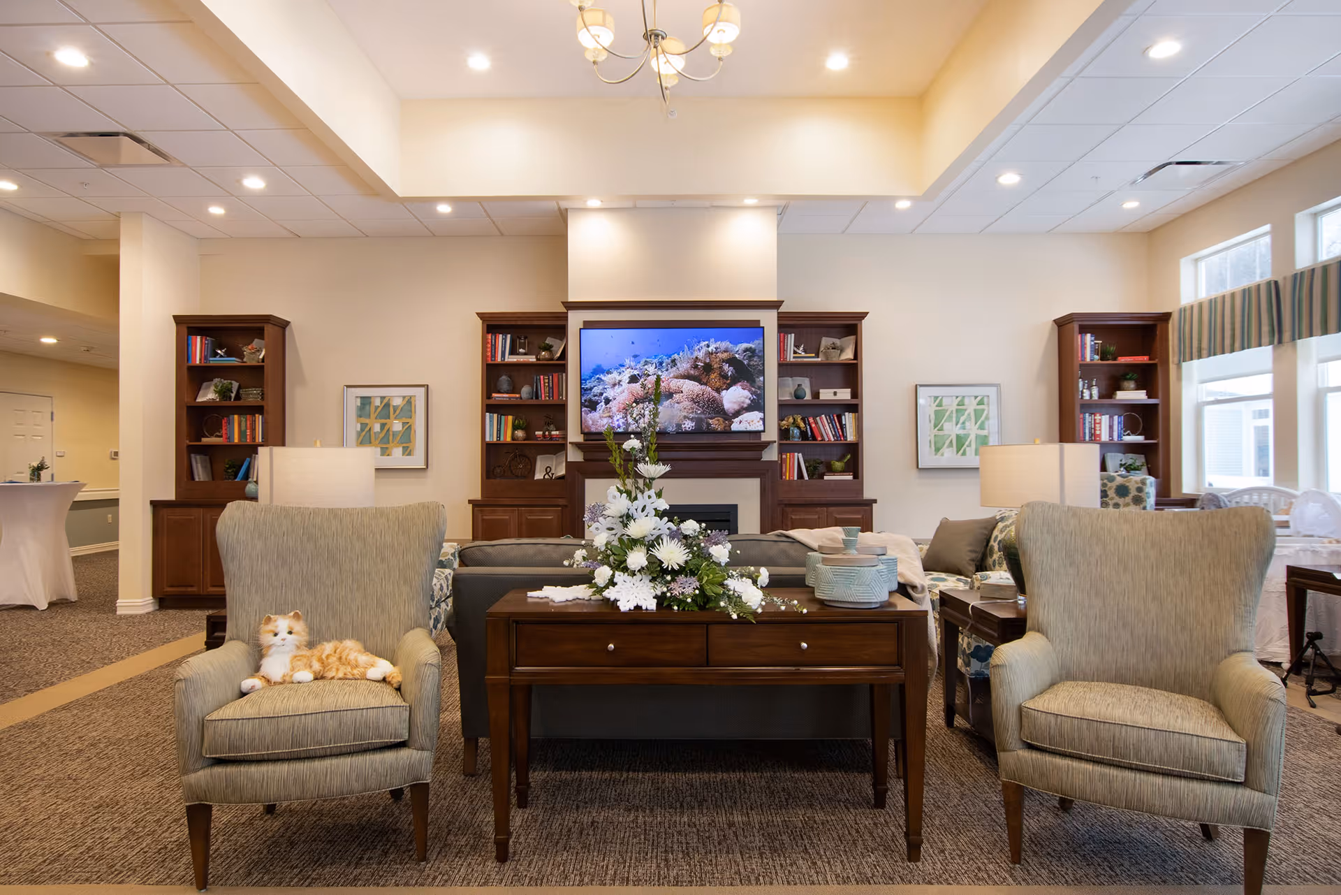 A bright communal living room with two armchairs flanking a console table topped with a floral arrangement, sofa, bookshelves and a television above a fireplace.