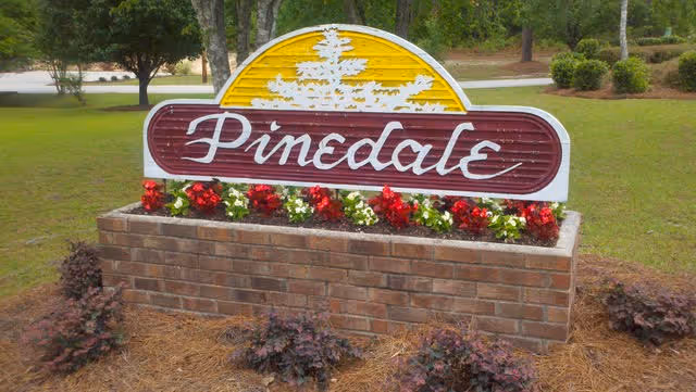 Wooden entrance sign reading "Pinedale" mounted on a brick planter with red and white flowers and lawn behind it.
