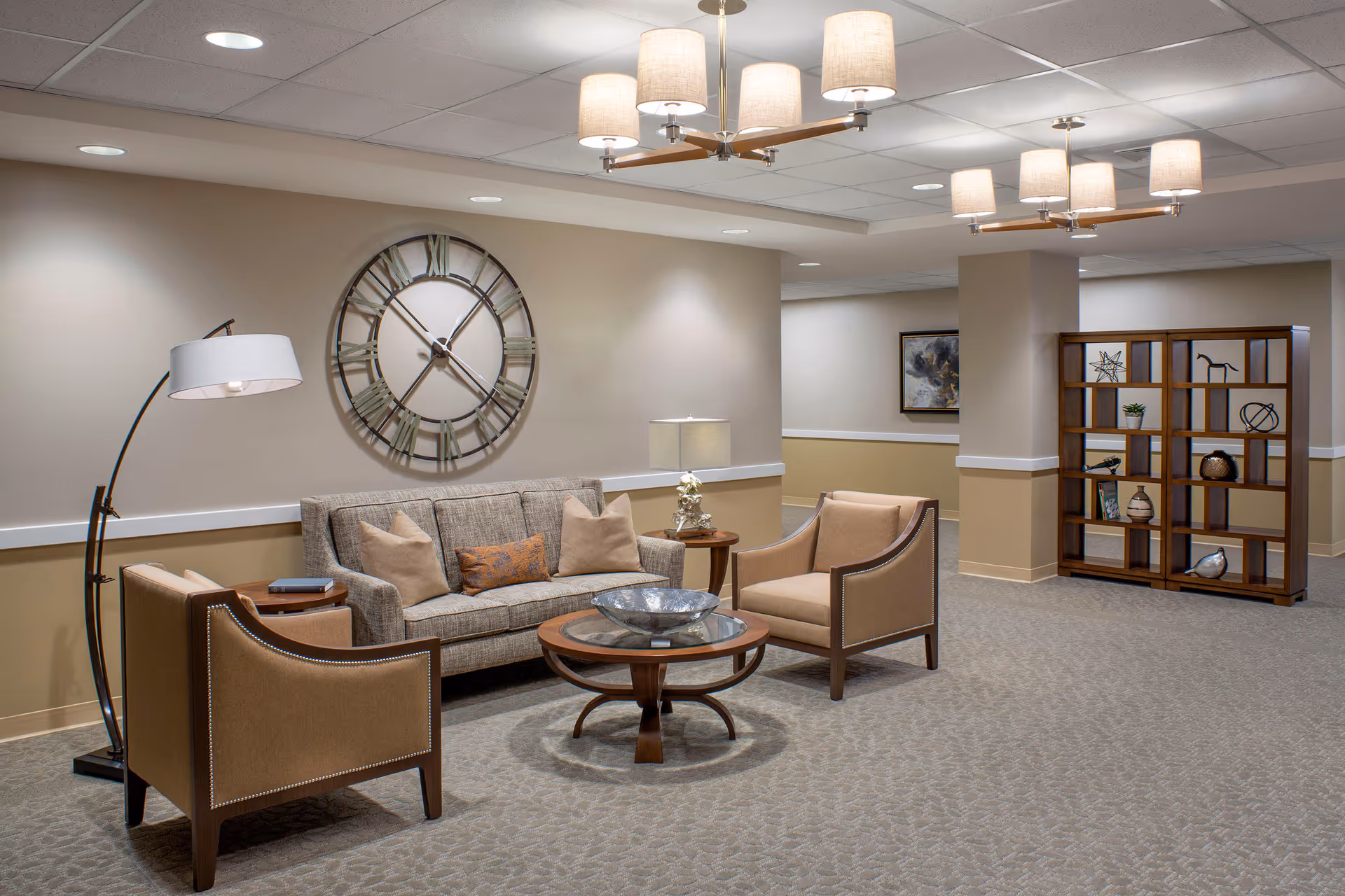 A furnished senior living lounge with a sofa, two armchairs, coffee tables, a large wall clock, and decorative shelving under soft ceiling lights.