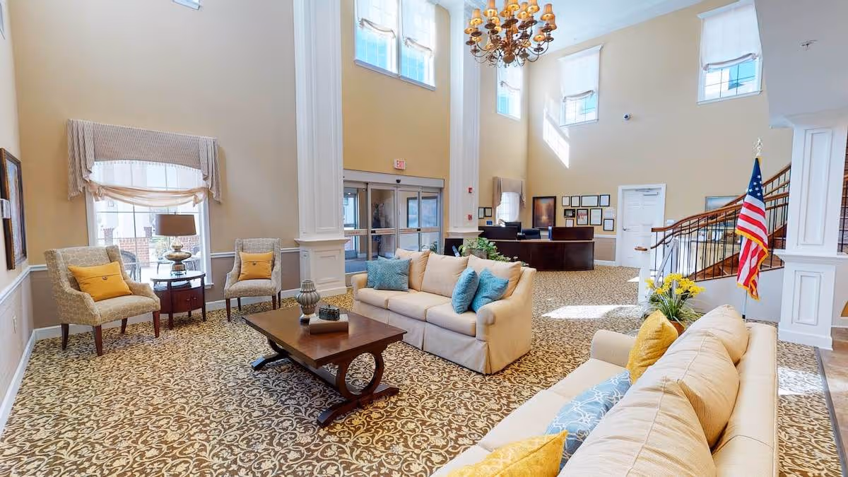 Sunlit high-ceilinged senior living lobby with sofas, armchairs, a coffee table, reception desk and an American flag.