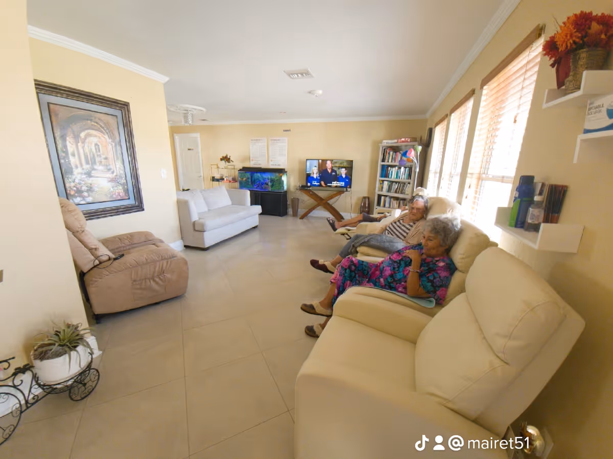 A bright living room in an assisted living facility with two elderly women sitting on cream-colored recliners near windows with blinds. The room has beige walls, a large painting on one wall, a beige recliner chair, a white loveseat, a fish tank, a TV on a wooden stand, and a bookshelf filled with books and decorations.