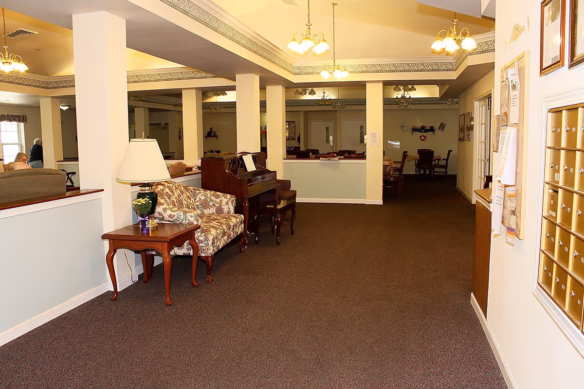 Carpeted communal lounge area with upholstered seating, a piano, side table and lamp in a senior living facility.