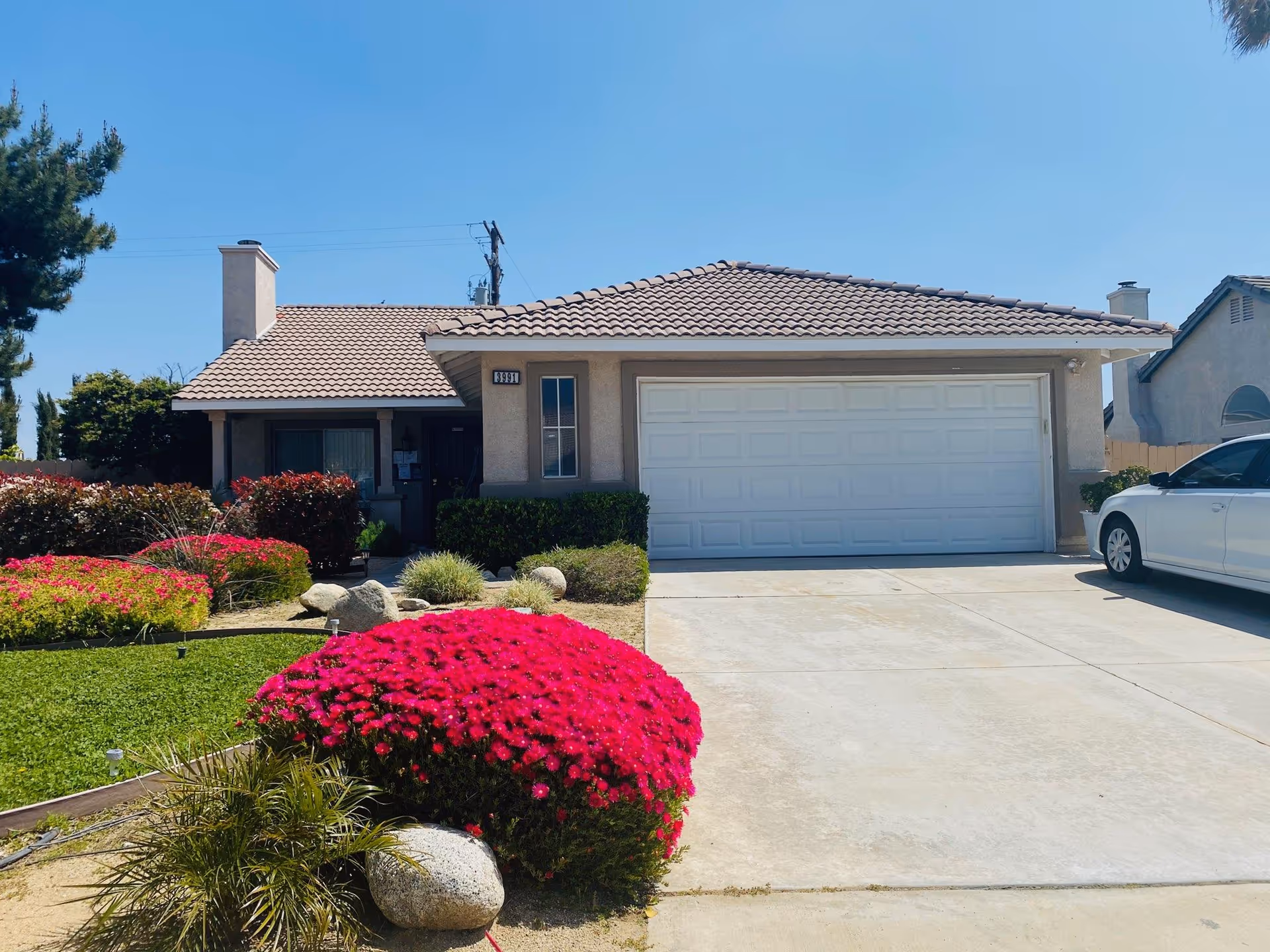 Front exterior view of a single-story house with a tiled roof, a two-car garage with a white door, a driveway with a white car parked on the right, and a well-maintained garden featuring bright pink flowers and green shrubs under a clear blue sky.