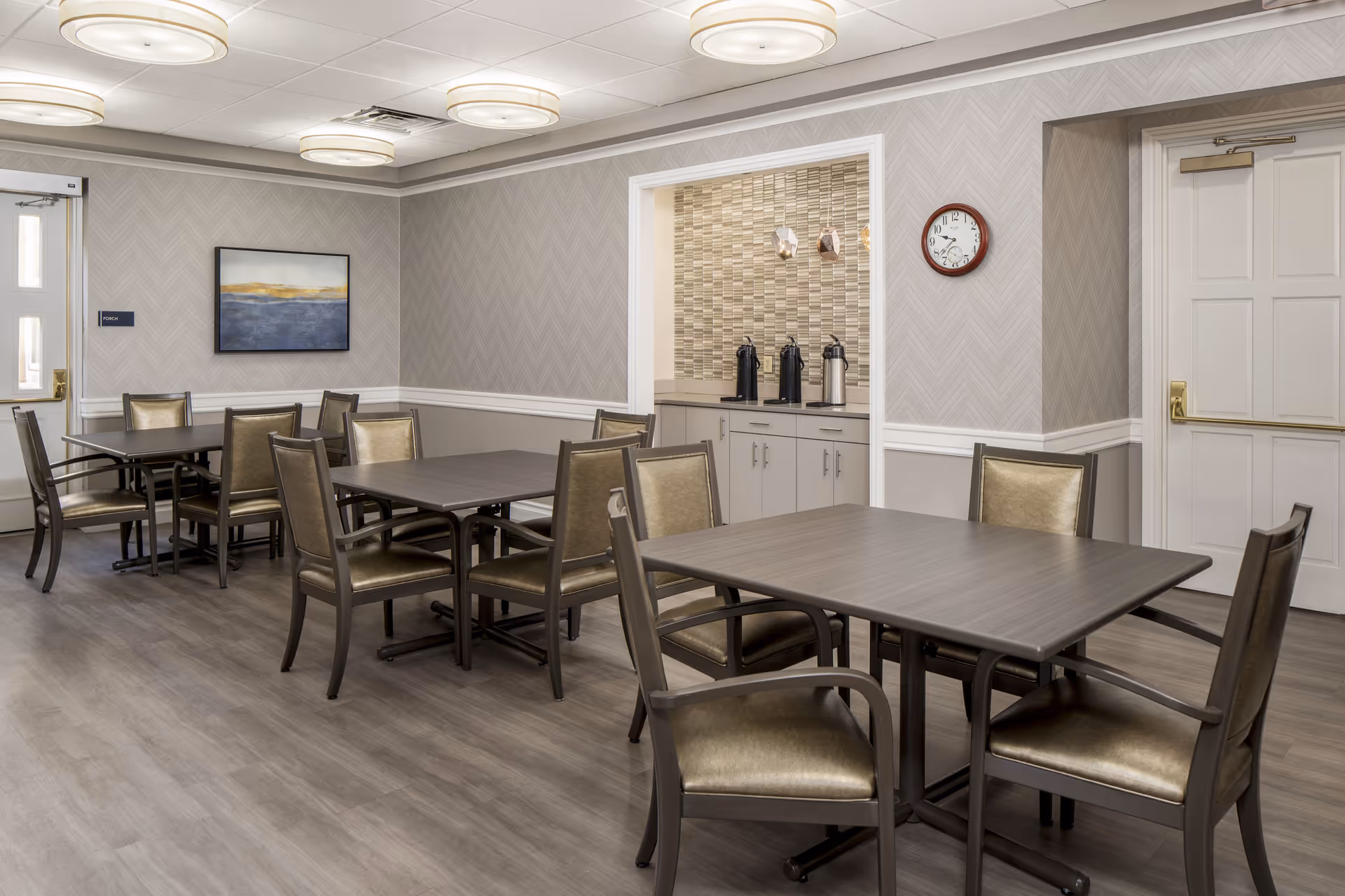 A dining room in an assisted living facility with several tables and chairs arranged neatly. The room features light gray walls with a chevron pattern, a wall clock, a framed abstract painting, and a small counter area with coffee dispensers. The floor is covered with wood-like vinyl flooring, and there are modern ceiling lights.