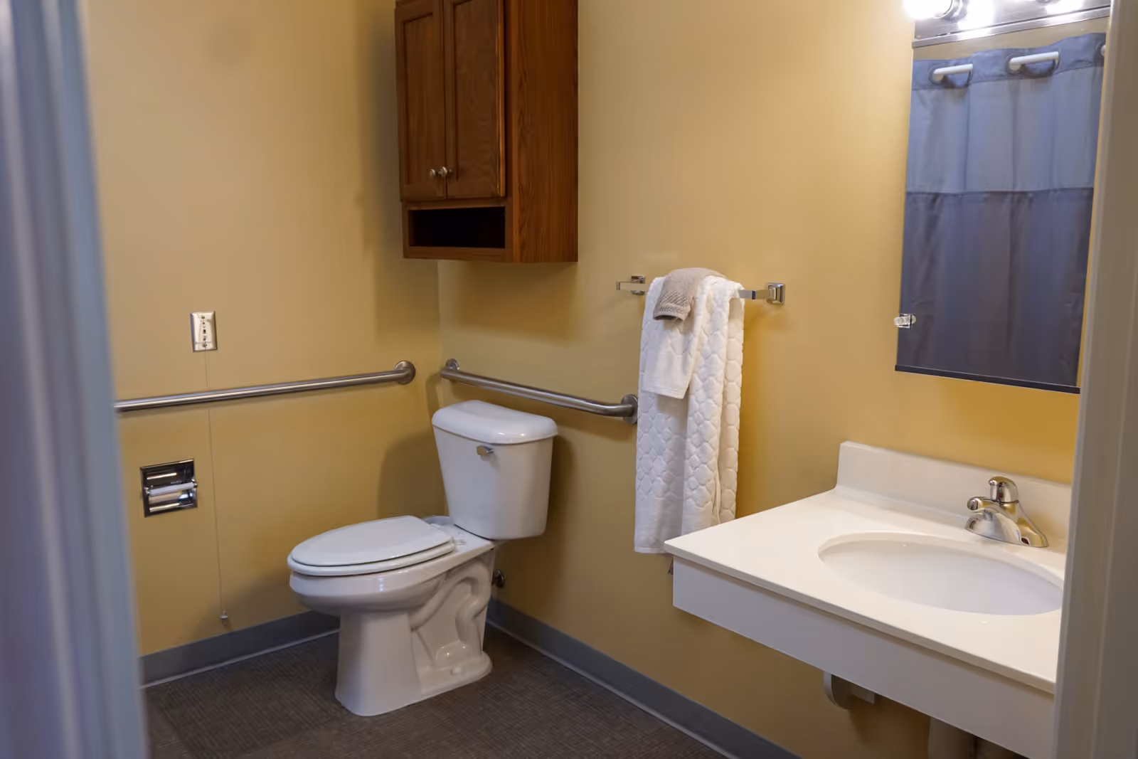 A clean and accessible bathroom with beige walls, a white toilet with grab bars on the walls, a white sink with a faucet, a mirror above the sink, a towel rack holding white towels, and a wooden cabinet mounted on the wall.