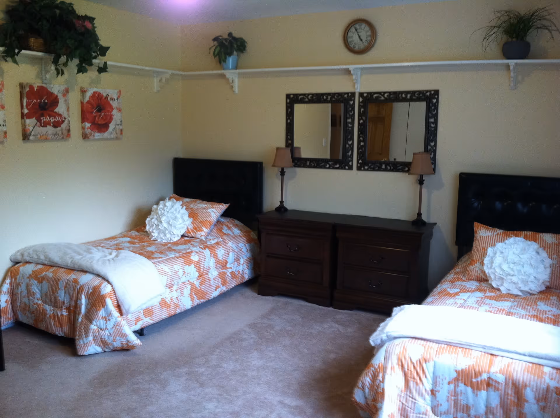 A bedroom with two twin beds featuring orange and white floral bedspreads, each bed has a white decorative pillow and a folded blanket at the foot. Between the beds is a dark wooden dresser with two matching table lamps and two framed mirrors above it. The walls are light-colored with a shelf running along the top, holding potted plants and decorative items. There are two floral paintings on the left wall and a clock above the mirrors.