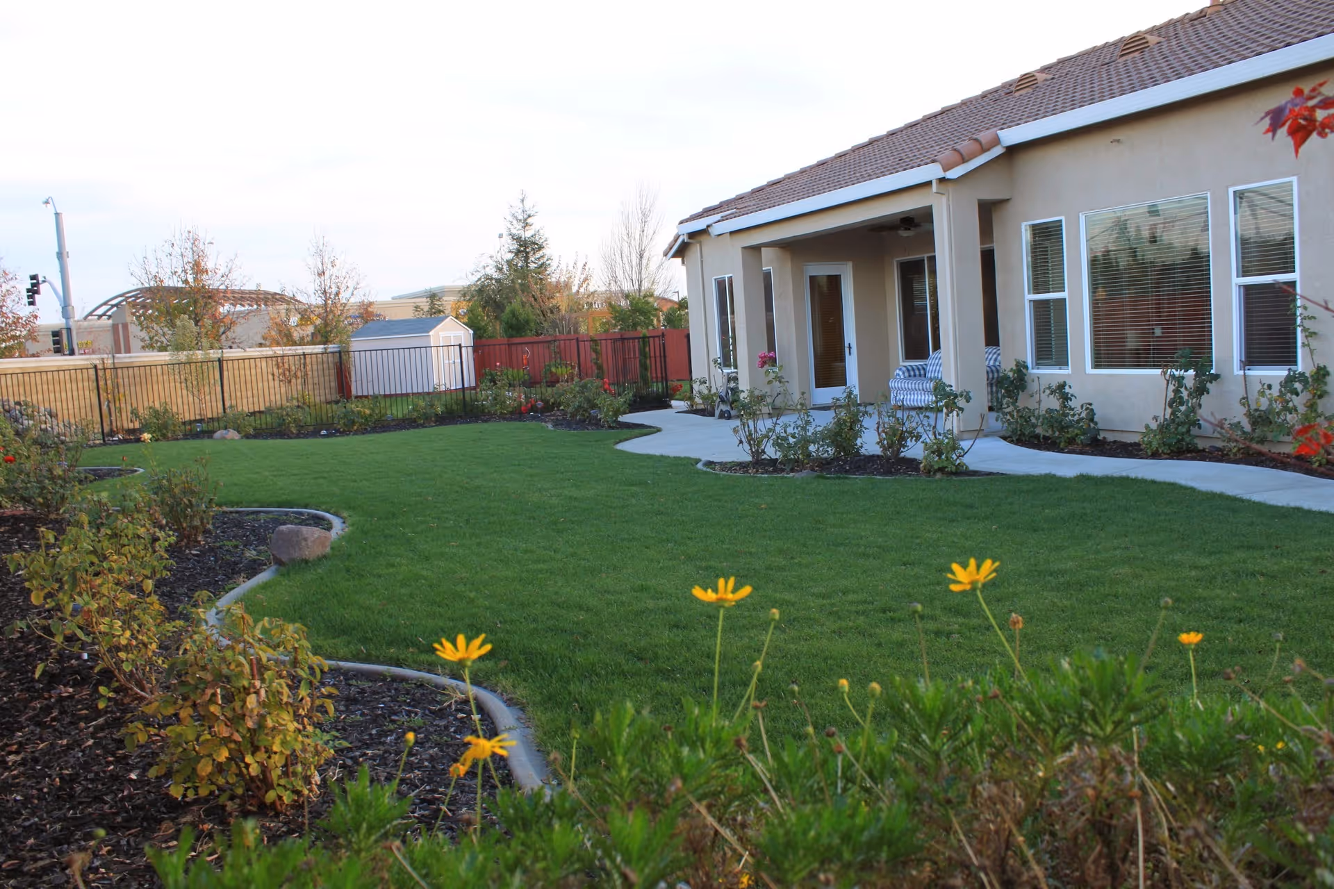 A single-story beige senior living facility with a curved walkway, green lawn, and landscaped garden beds along the exterior.
