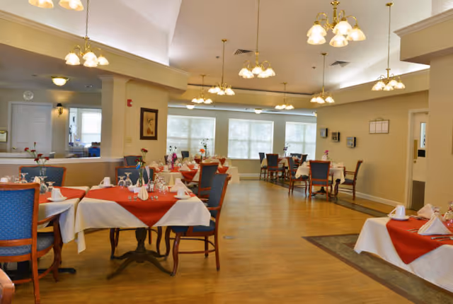 A spacious dining room with multiple tables covered in white tablecloths and red table runners. Each table is set with napkins, glasses, and small flower vases. The room has wooden flooring, blue cushioned chairs, and several ceiling light fixtures. Large windows allow natural light to fill the space.